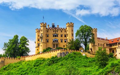 Hohenschwangau Castle surrounded by lush greenery under a blue sky.