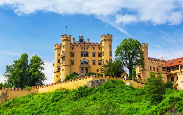 Hohenschwangau Castle surrounded by lush greenery under a blue sky.
