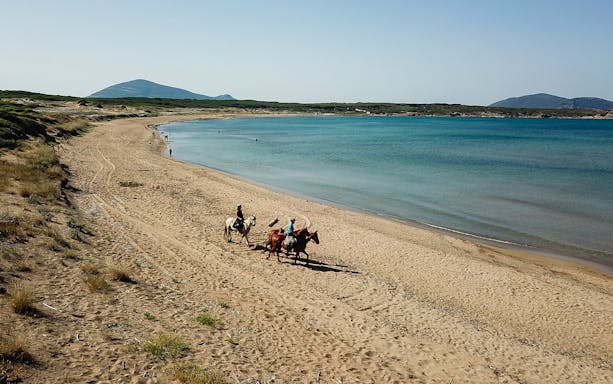 Horseback riders on Porto Ferro beach with distant hills and blue sea.