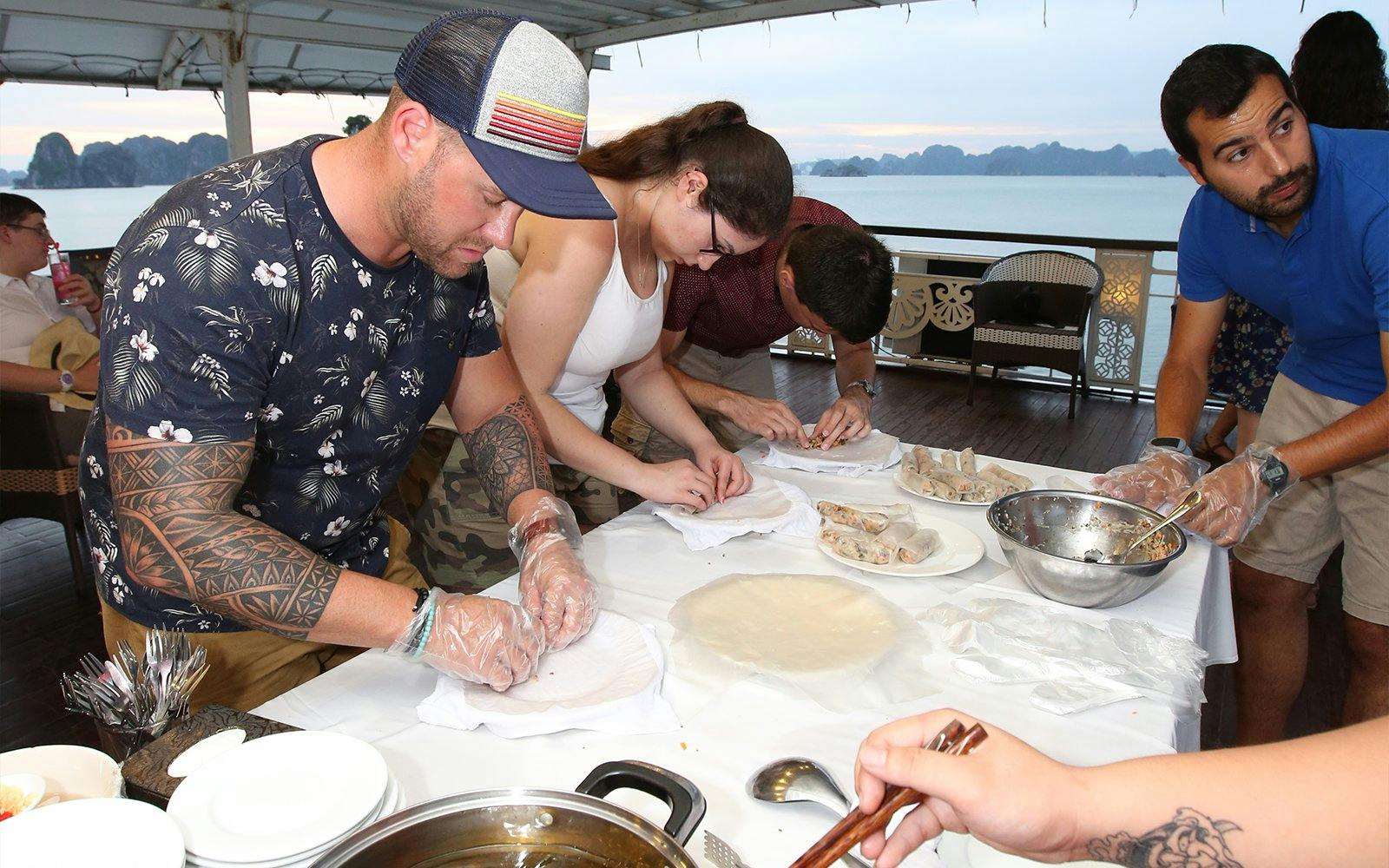 Tourists making spring rolls on a Halong Bay cruise with scenic views in the background.