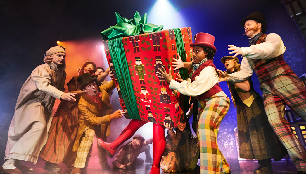 Actors in Victorian costumes struggle with a giant gift box on stage during "Christmas Carol Goes Wrong" in Sydney.