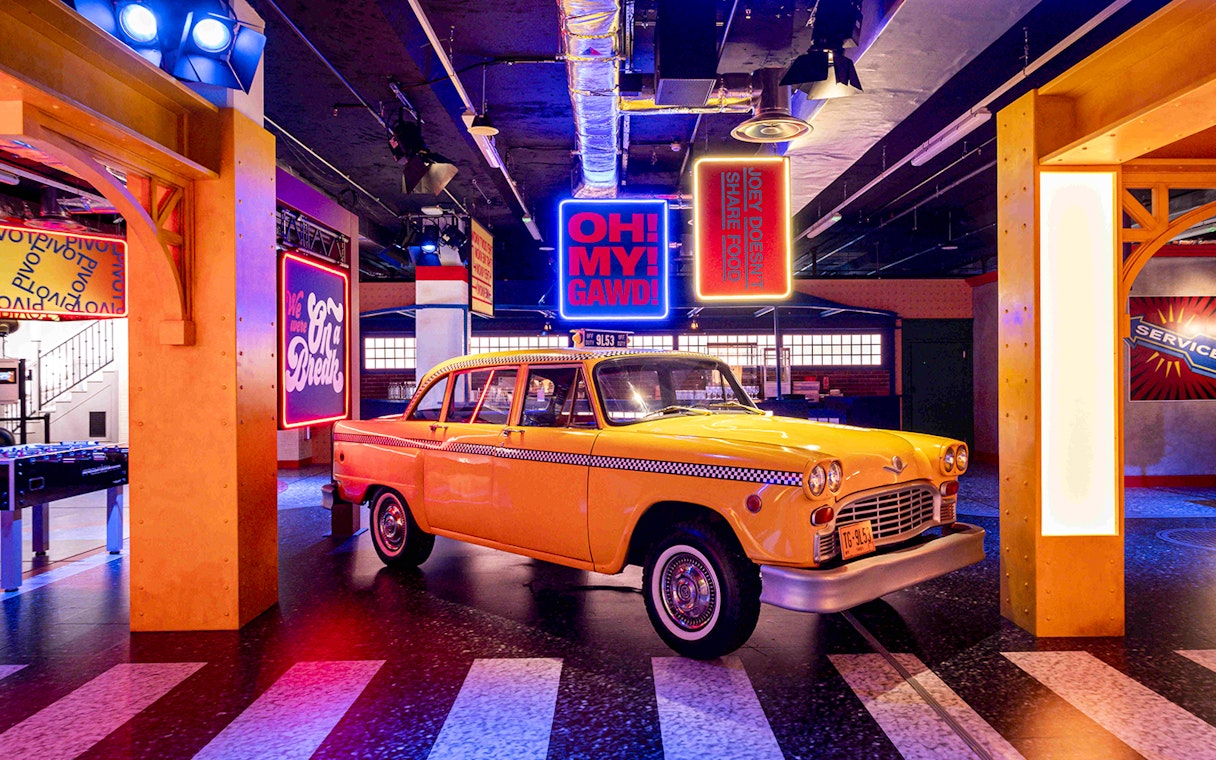 Yellow taxi cab at The FRIENDS™ Experience with iconic show quotes in neon signs.