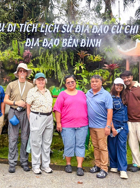 Tourists posing at Cu Chi Tunnels entrance, Vietnam, with lush greenery and signage in the background.