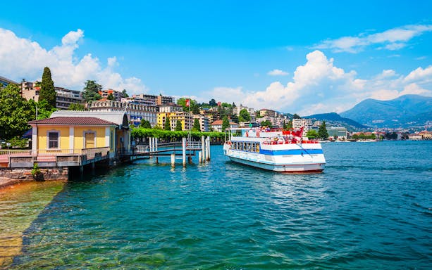 Cruise boat on Lake Lugano with cityscape and mountains in the background.