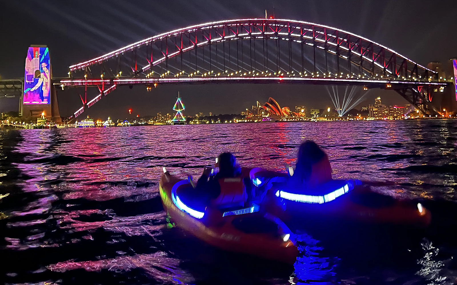 Kayakers paddle under Sydney Harbour Bridge with city skyline and fireworks at night.