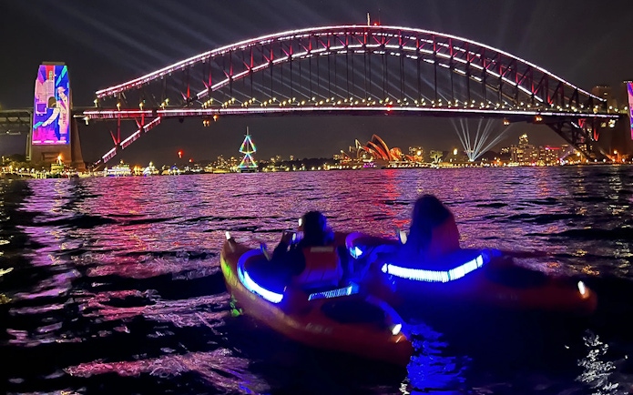 Kayakers paddle under Sydney Harbour Bridge with city skyline and fireworks at night.