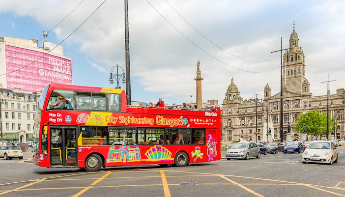 Open-top sightseeing bus in Glasgow city center near historic buildings.