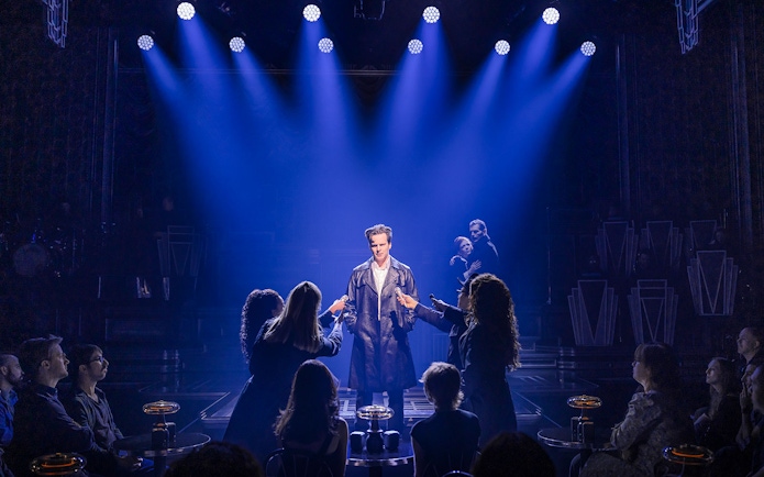 Performer on stage under blue lights with audience in a theater setting.