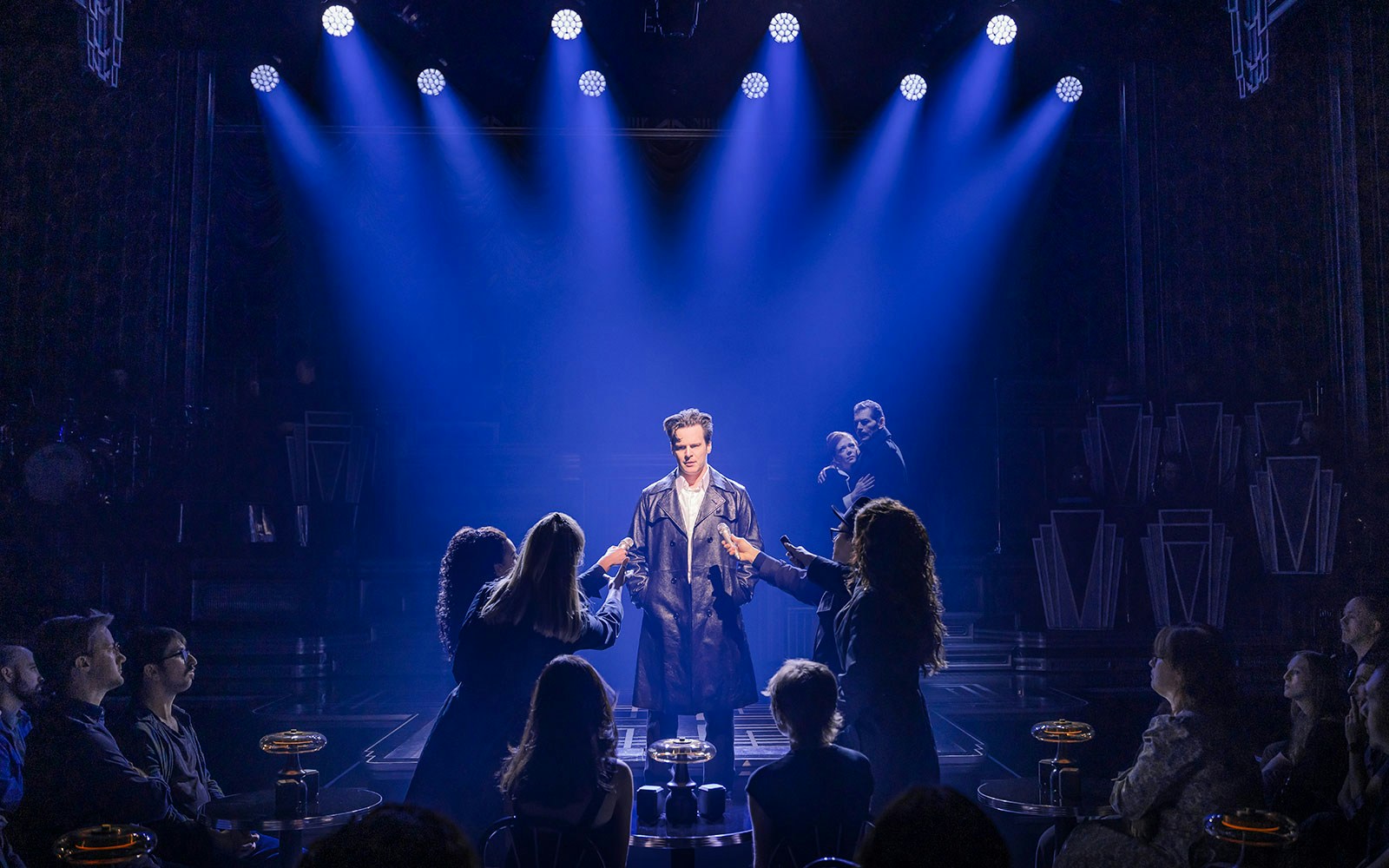 Performer on stage under blue lights with audience in a theater setting.