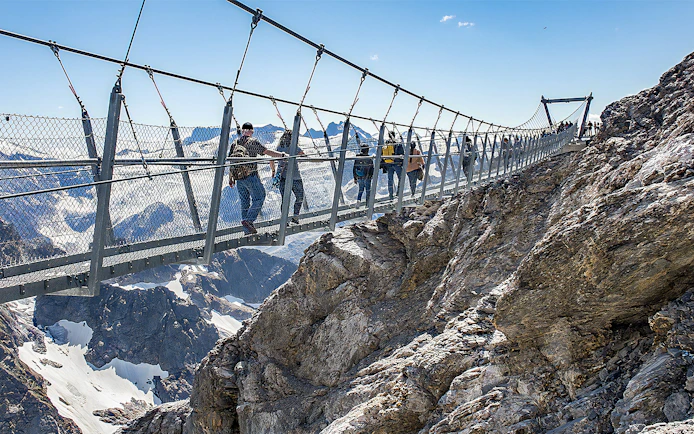 Tourists crossing a suspension bridge at Mt. Titlis with snowy peaks in the background.