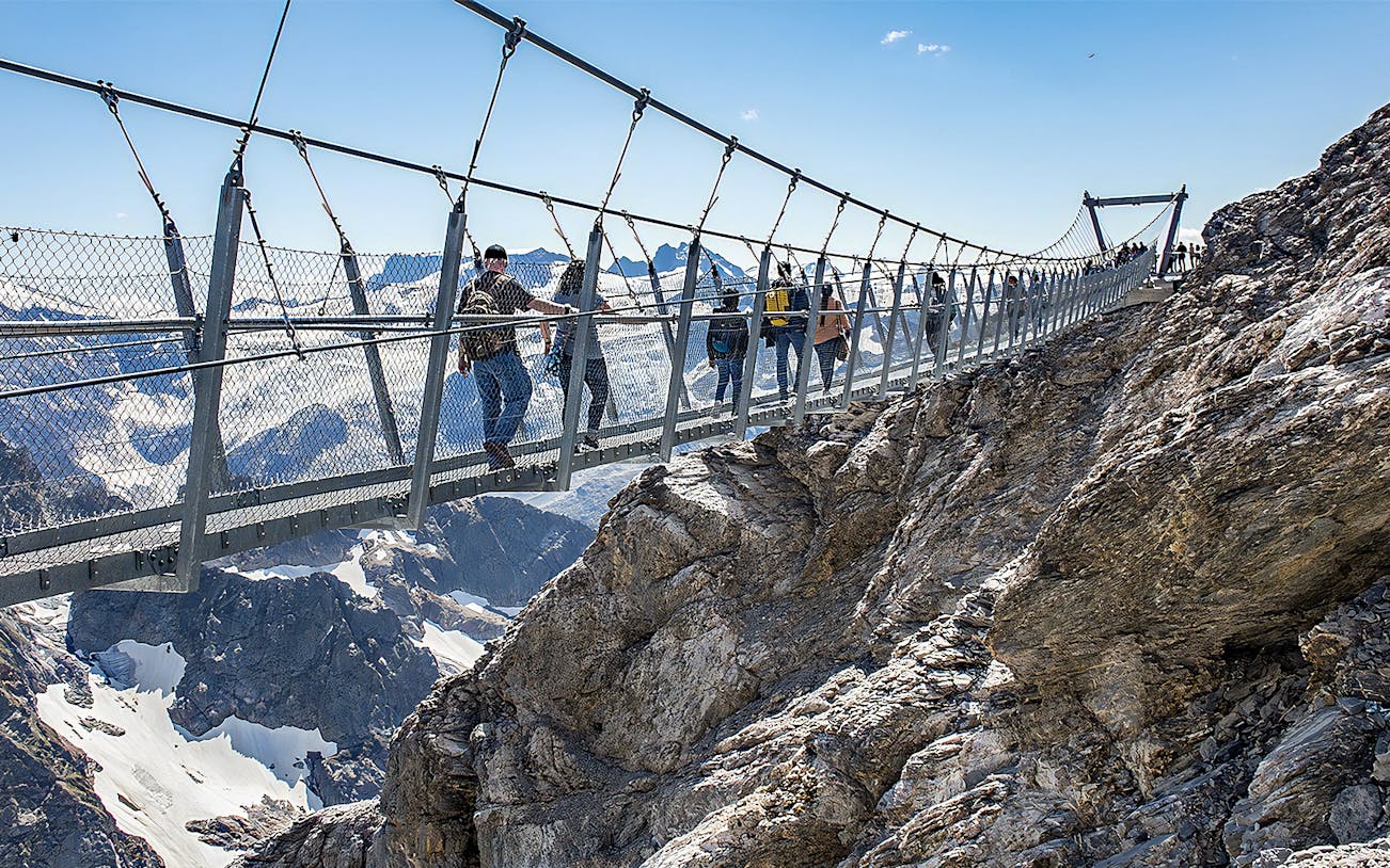 Tourists crossing a suspension bridge at Mt. Titlis with snowy peaks in the background.