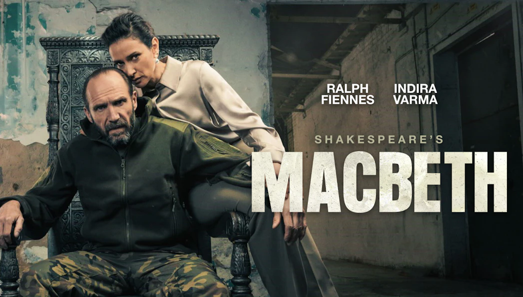 Actors in a dramatic scene from Shakespeare's Macbeth, with a rustic backdrop.