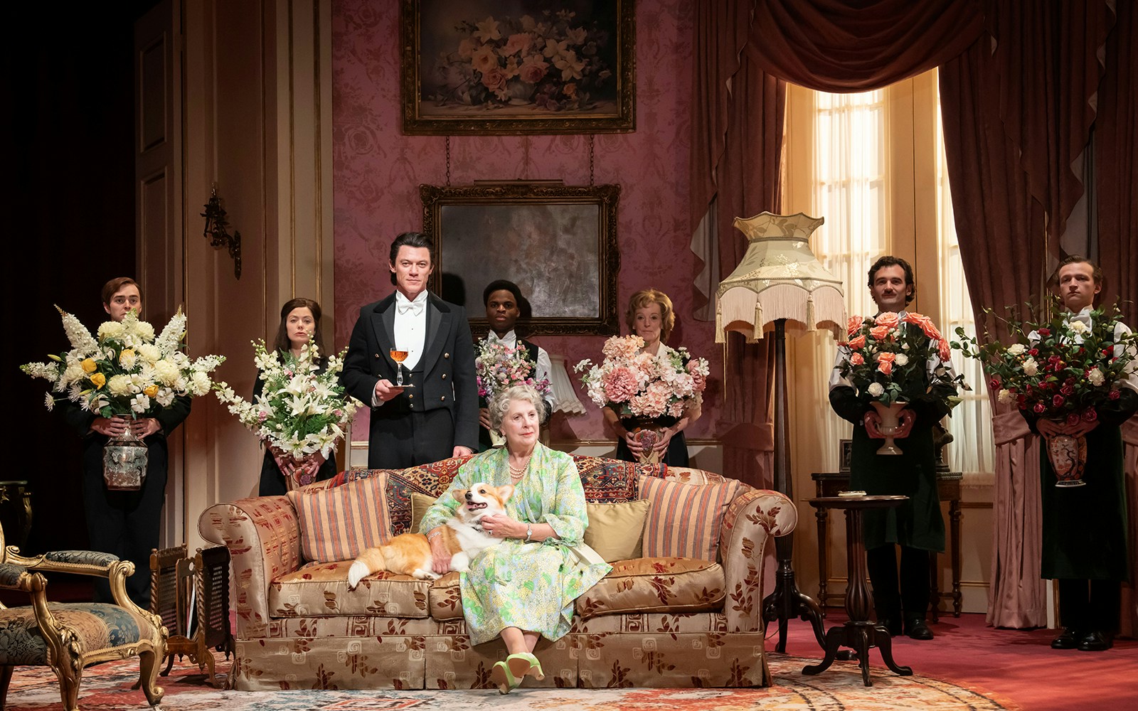 Elegant room with a woman on a sofa holding a dog, surrounded by people with flower arrangements, related to Backstairs Billy.