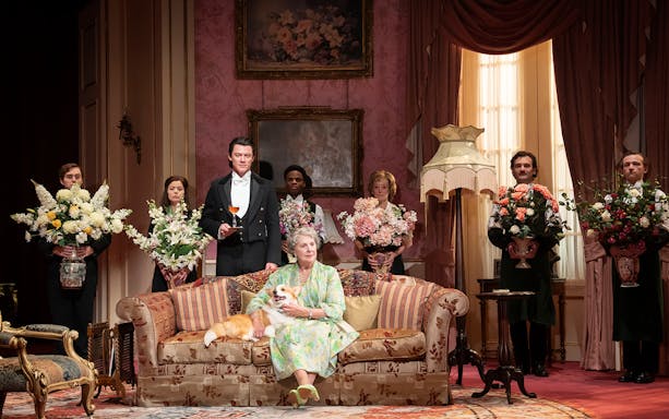 Elegant room with a woman on a sofa holding a dog, surrounded by people with flower arrangements, related to Backstairs Billy.