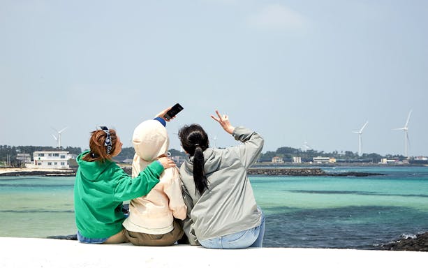 Three people taking a selfie by the sea with wind turbines in the background in Korea.