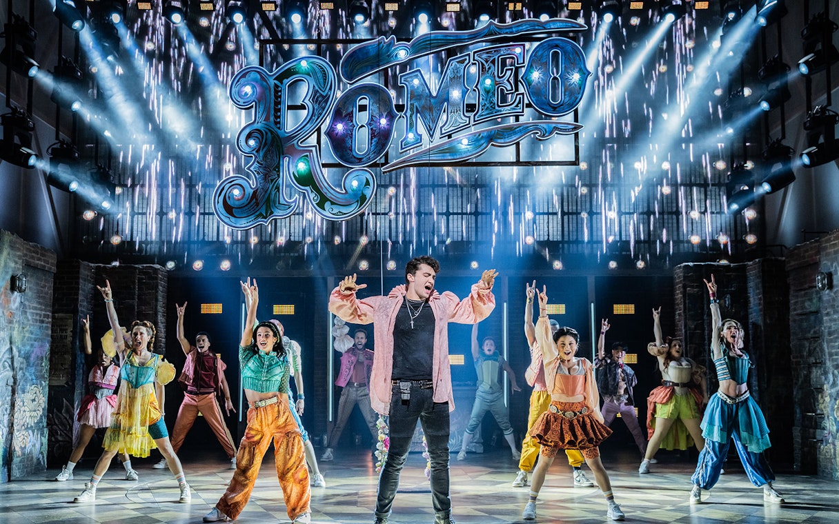 Performers on stage during a scene from the musical "& Juliet" with a "Romeo" sign above.