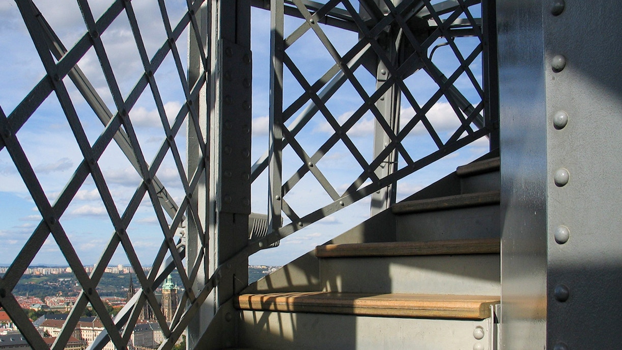 Stairs inside Petrin Tower with a view of Prague through metal lattice.
