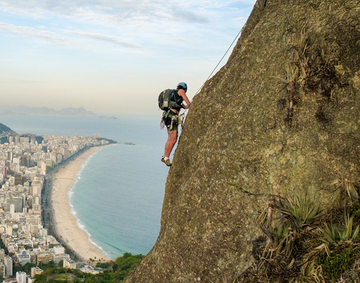 Climber scaling rock wall with Rio de Janeiro's Copacabana Beach in the background.