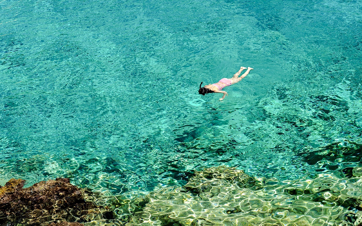 Woman snorkeling in clear turquoise water near a rocky reef.