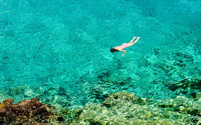 Woman snorkeling in clear turquoise water near a rocky reef.