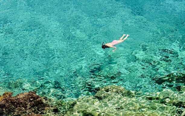 Woman snorkeling in clear turquoise water near a rocky reef.