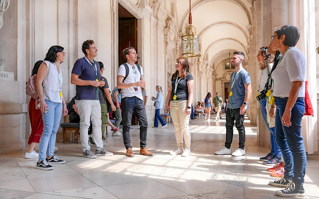 Group on a guided tour at the Prado Museum, Madrid, listening to a guide in a sunlit hallway.