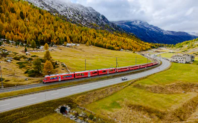 Red Bernina train traveling through Swiss Alps landscape near St. Moritz, surrounded by autumn trees.