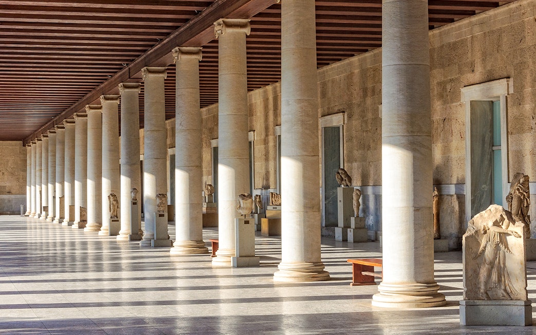 Ancient Agora of Athens colonnade with statues and sunlight streaming through columns.