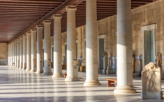 Ancient Agora of Athens colonnade with statues and sunlight streaming through columns.