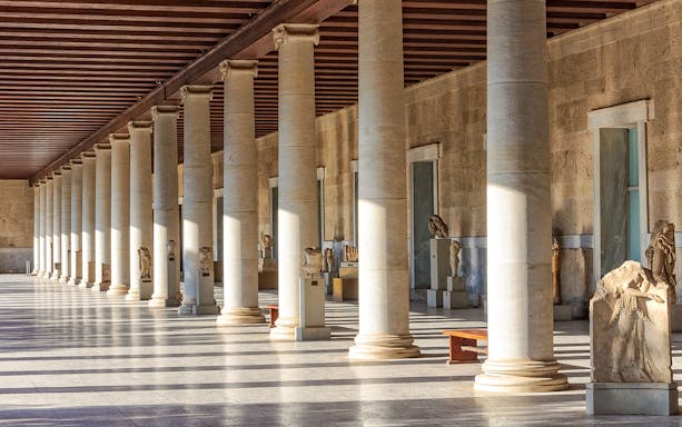 Ancient Agora of Athens colonnade with statues and sunlight streaming through columns.