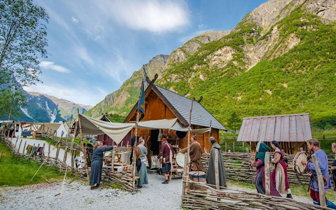 Viking reenactors in traditional attire inside a Viking Village with wooden huts and mountainous backdrop.