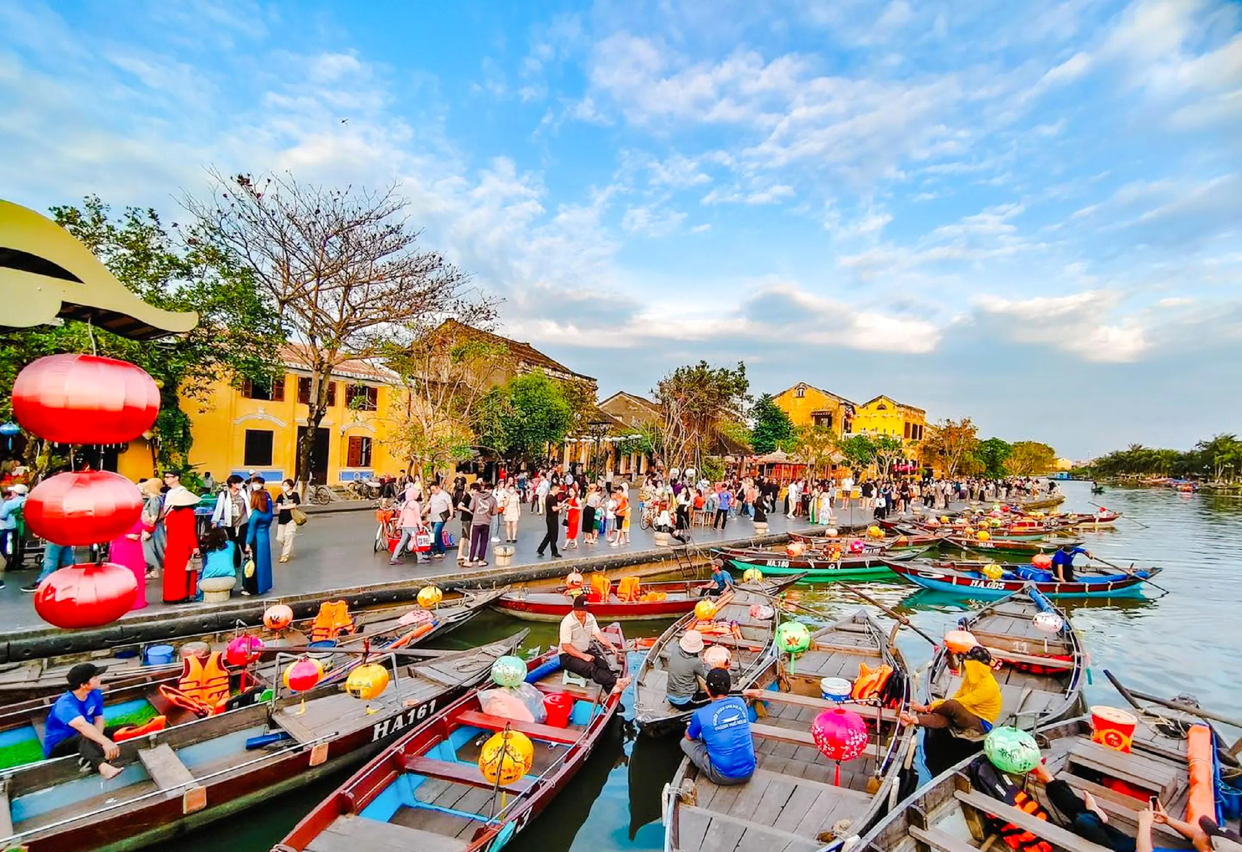 Boats with lanterns on the river in Hoi An, Vietnam, with people walking along the waterfront.