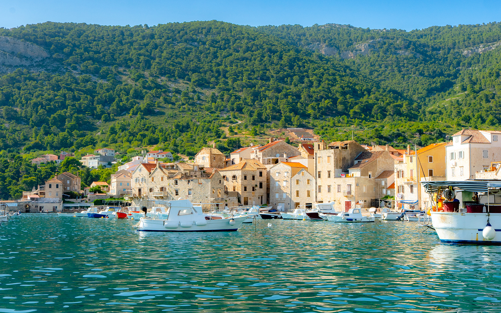 Boats docked at Komiža harbor with stone buildings and green hills in the background.
