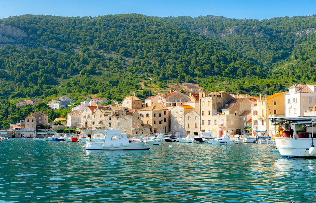 Boats docked at Komiža harbor with stone buildings and green hills in the background.