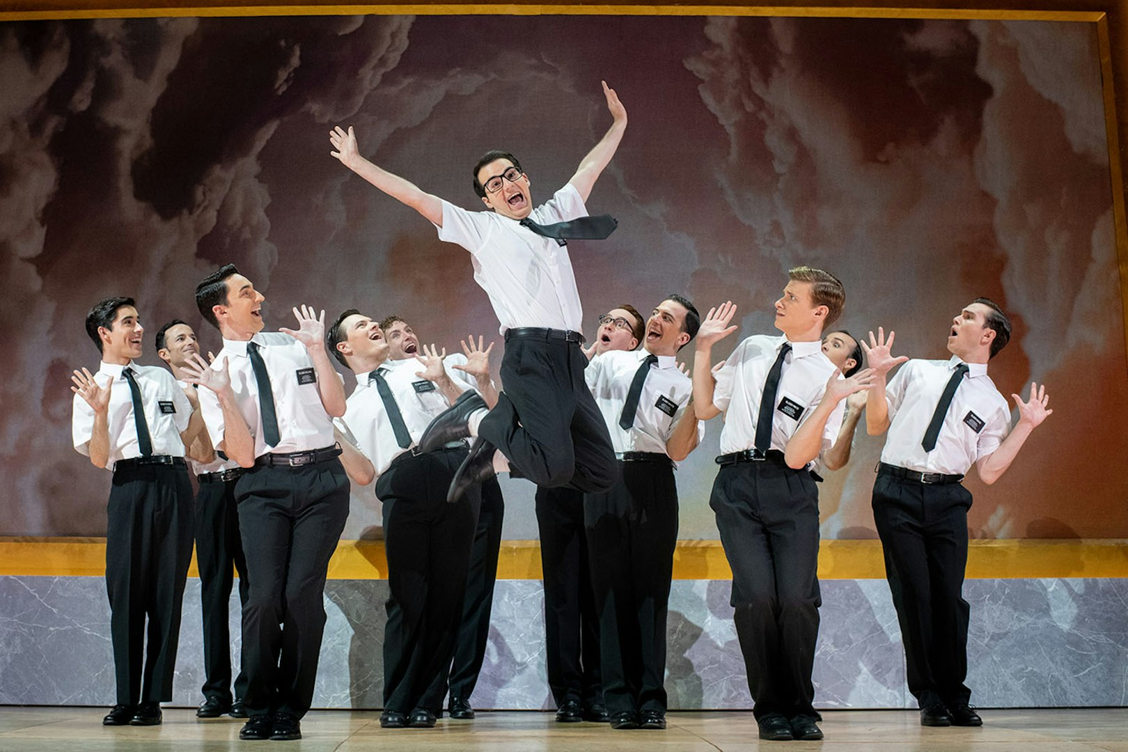 Performers in "The Book of Mormon" musical on stage, one jumping energetically.