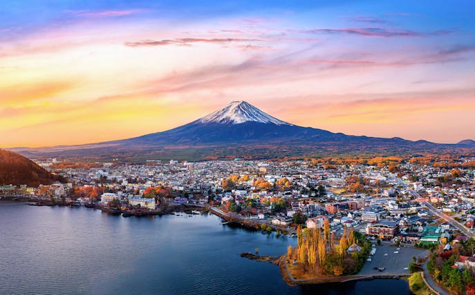 Aerial view of Mount Fuji with a cityscape and lake in the foreground at sunset.