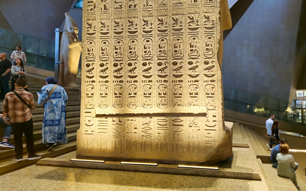 Hieroglyphs on stone slab at Grand Egyptian Museum, Cairo, with visitors exploring the exhibit.