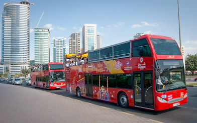 Red double-decker bus on Sharjah Hop-On-Hop-Off tour with city skyscrapers in the background.