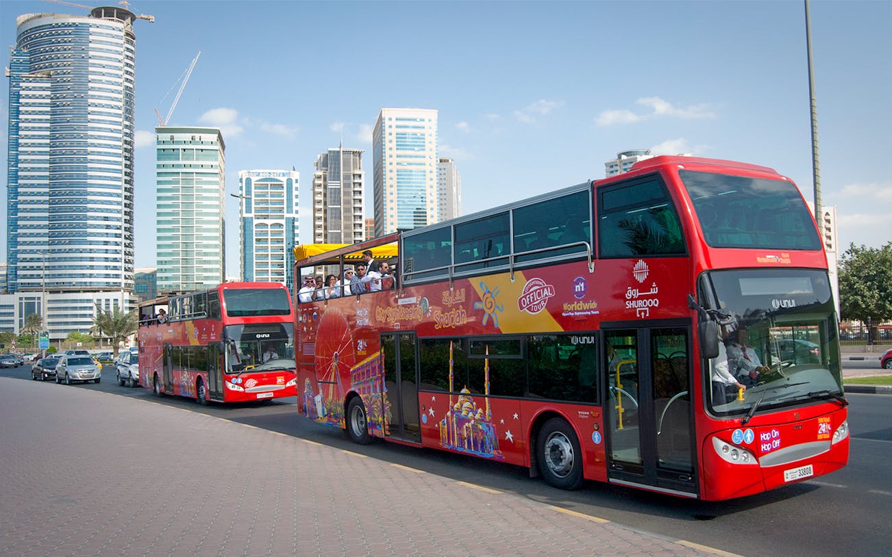 Red double-decker bus on Sharjah Hop-On-Hop-Off tour with city skyscrapers in the background.