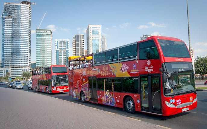 Red double-decker bus on Sharjah Hop-On-Hop-Off tour with city skyscrapers in the background.