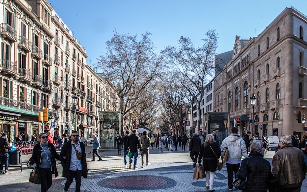 People walking along Las Ramblas in Barcelona, with historic buildings and trees lining the street.