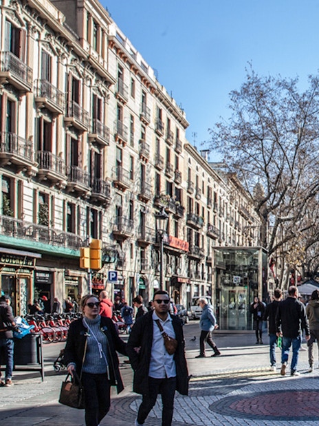 People walking along Las Ramblas in Barcelona, with historic buildings and trees lining the street.