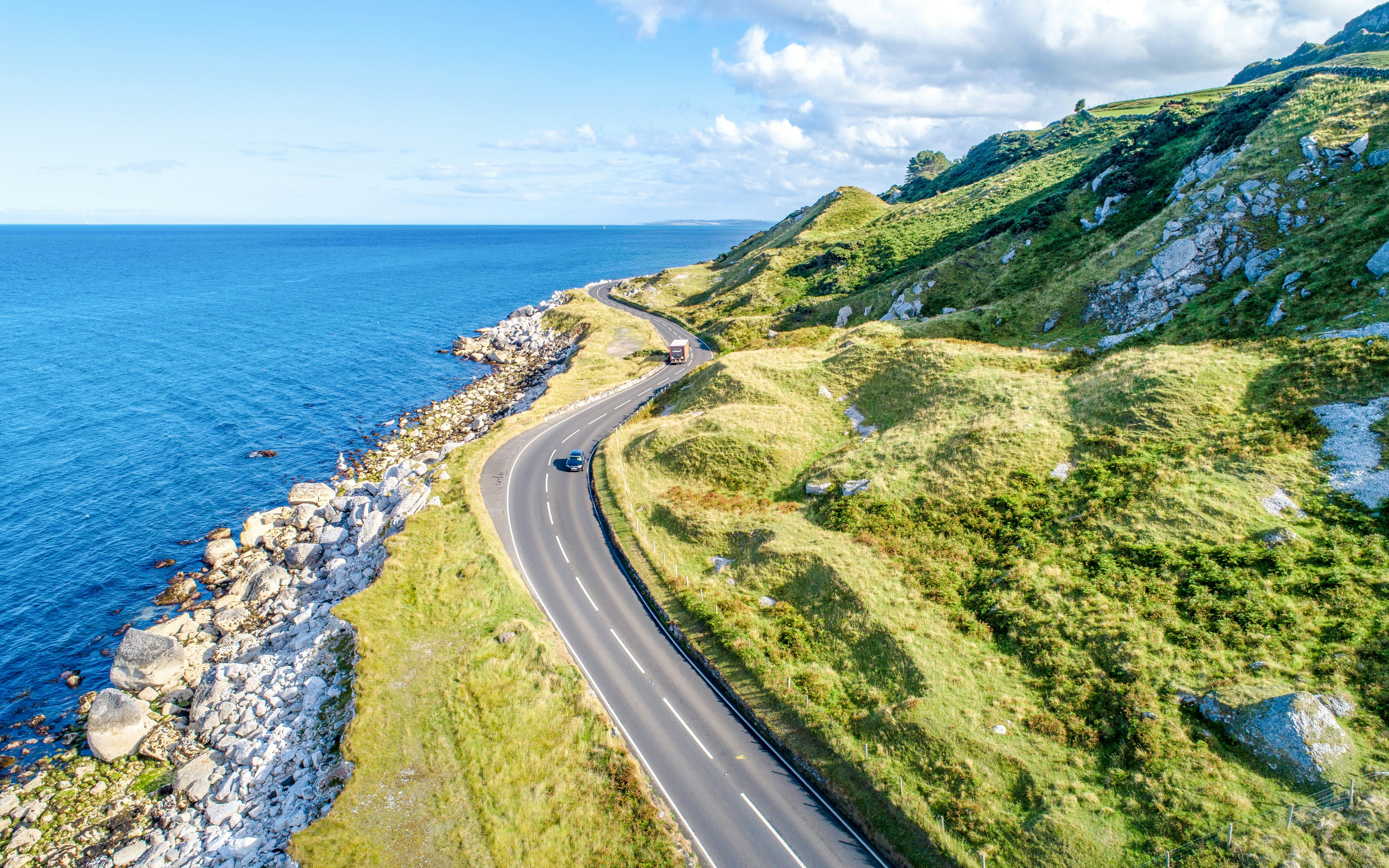 Antrim Coast Road winding along the rocky shoreline with green hills and blue sea.