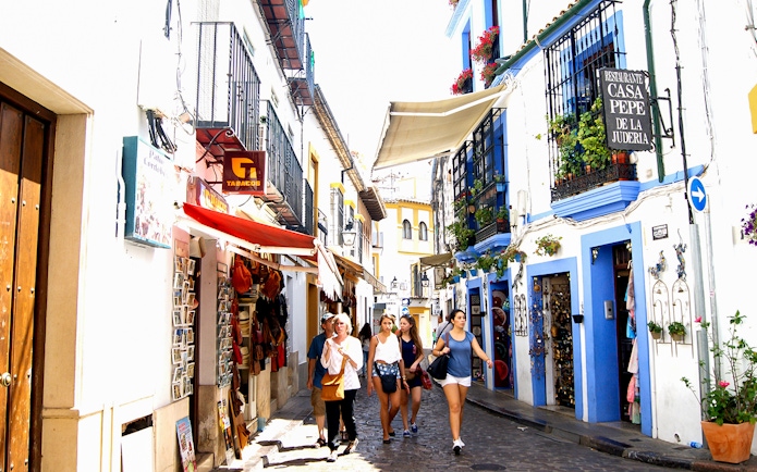 Street in Cordoba's Jewish Quarter with tourists walking, featuring colorful buildings and shops.