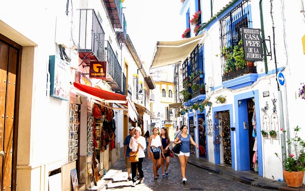 Street in Cordoba's Jewish Quarter with tourists walking, featuring colorful buildings and shops.
