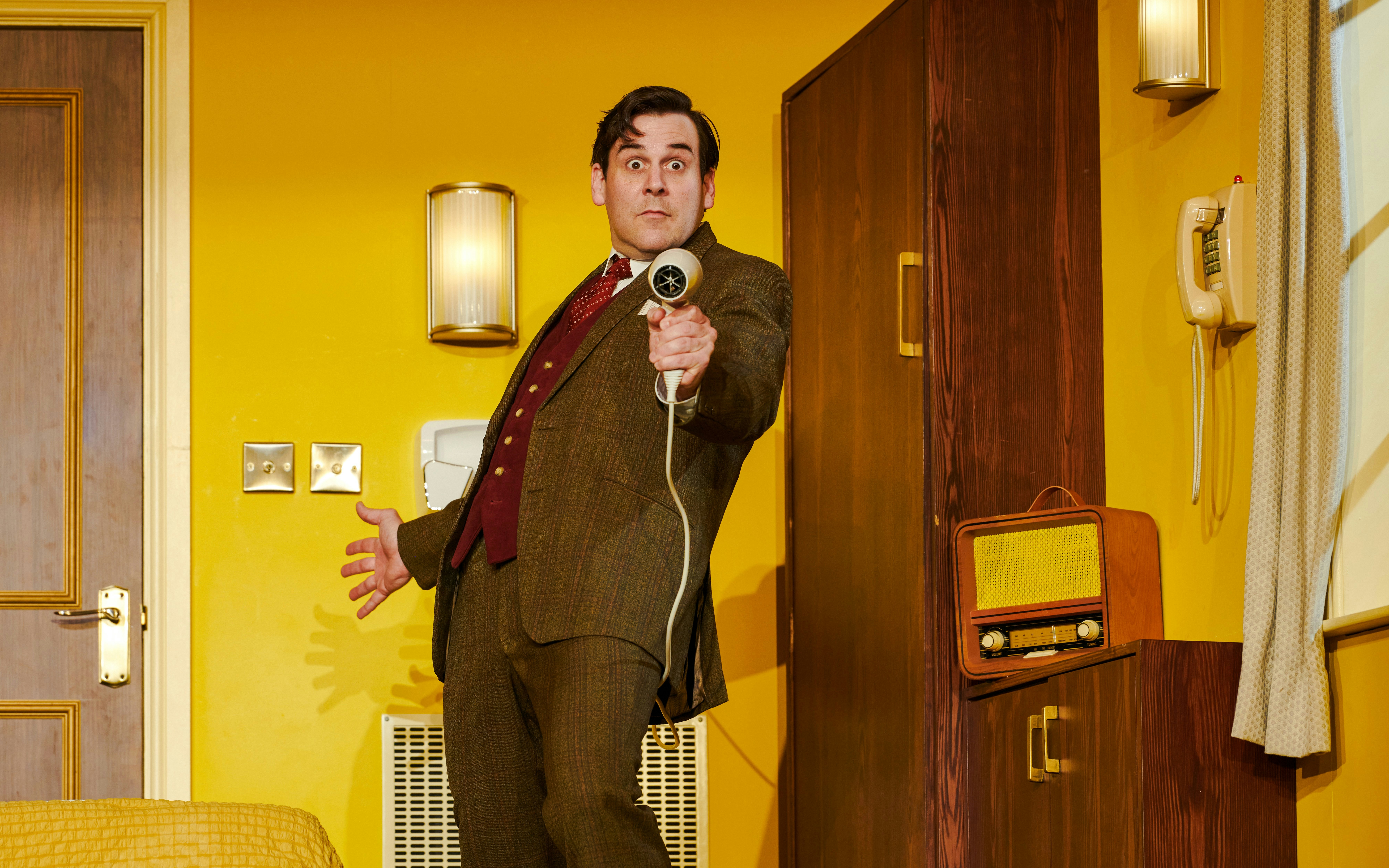 Man in suit holding a hairdryer like a gun in a comedic spy scene.