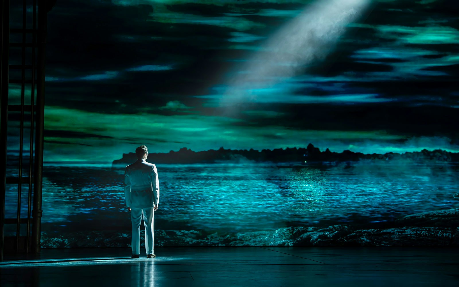 Man in white suit stands on stage with ocean backdrop in The Great Gatsby Musical.