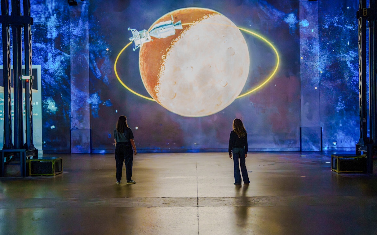 Visitors viewing a moon and spacecraft projection at The Moonwalkers Exhibition, Atelier des Lumières.