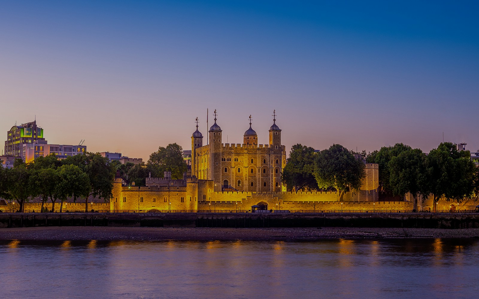 Tower of London illuminated at dusk, viewed from across the River Thames.