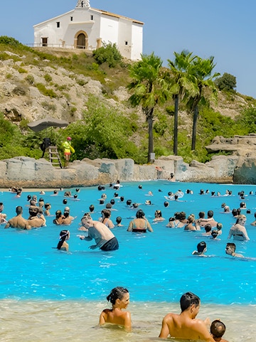 Visitors enjoying the wave pool at Aquopolis Cullera water park, Spain, with a hillside chapel in the background.